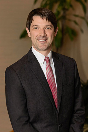 Headshot of a smiling man with short brown hair wearing a white shirt, red patterned tie, and black jacket.