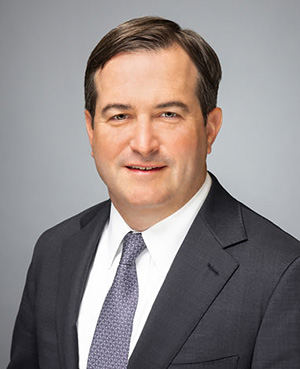Headshot of a man with short brown hair wearing a white shirt, dark blue patterned tie, and dark gray jacket.