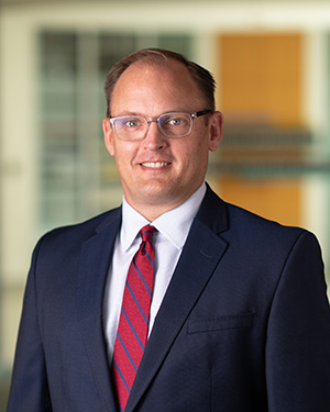 Headshot of a smiling man with balding light brown hair wearing light-colored glasses, a white shirt, a red and blue striped tie, and a navy jacket.