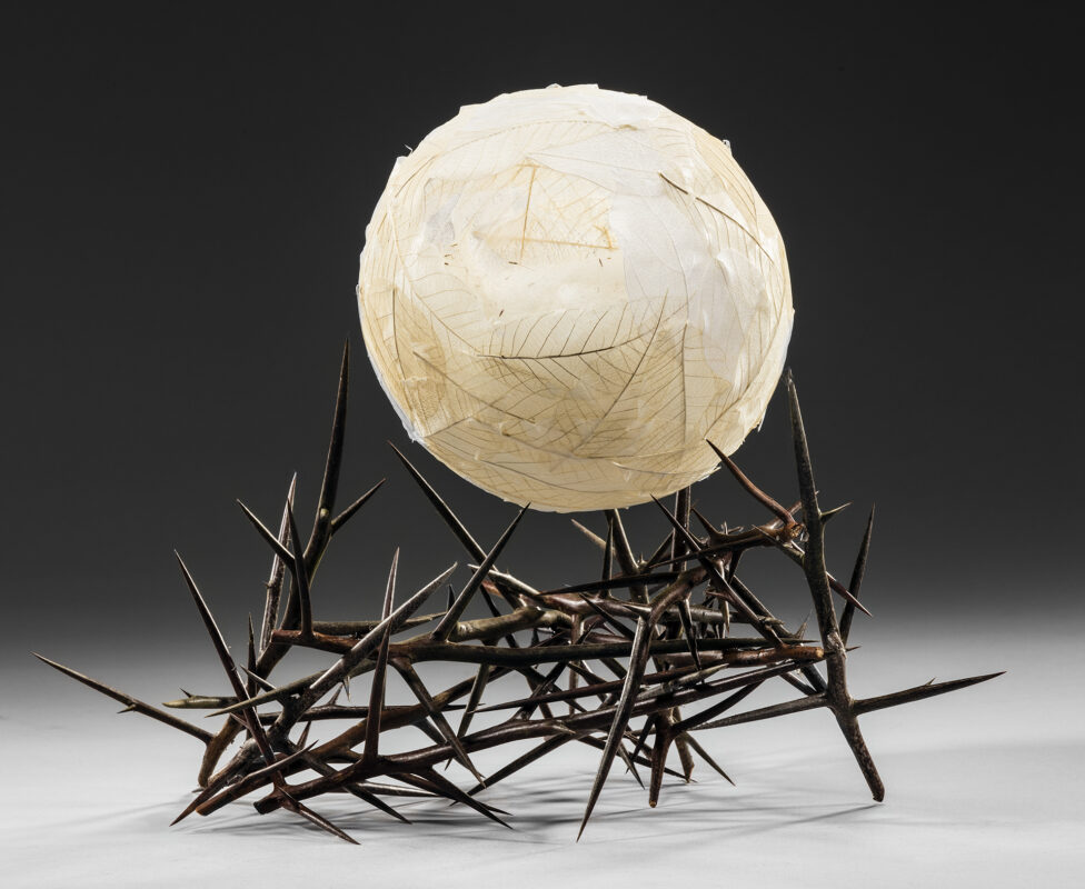 Photo of a sculpture comprised of a round white ball covered in faux leaves sitting on a bed on large metal thorns.