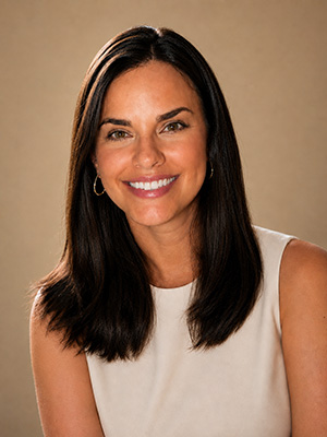 Headshot of a smiling woman iwth long brown hair wearing a cream colored sleeveless blouse and gold hoop earrings.