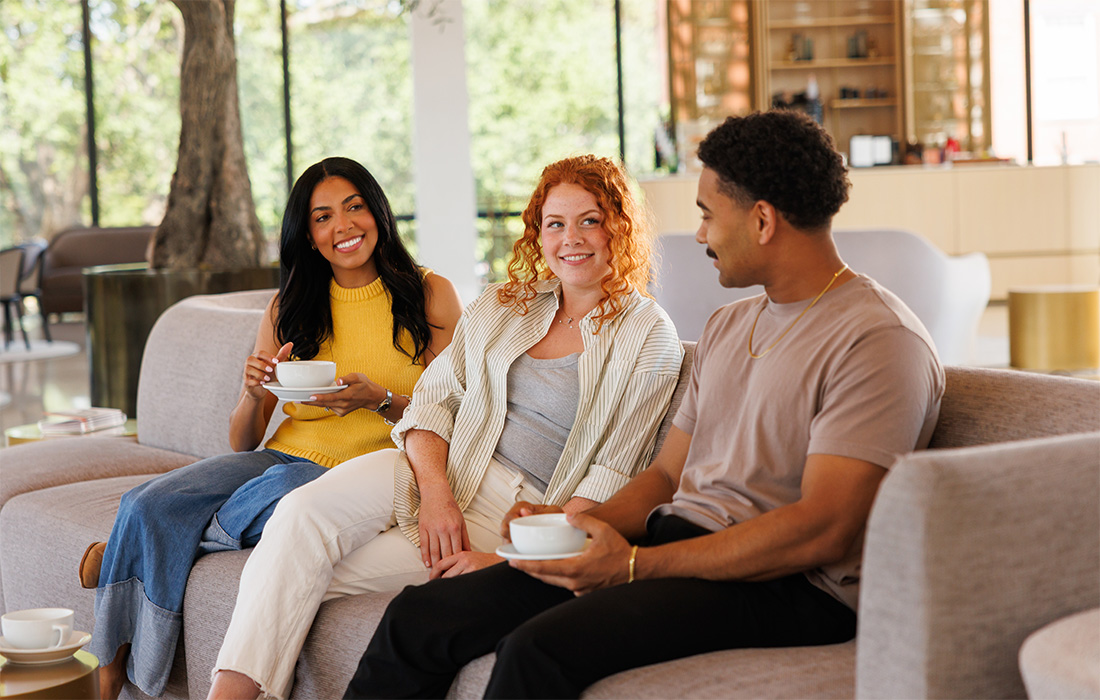 Photo of two women and a man smiling and holding white coffee cups while sitting on a couch in the Cultural Living Room at AMFA.
