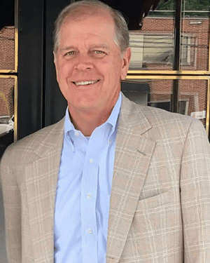 Photo of a smiling man with balding gray hair wearing a light blue shirt and a beige, checker-patterned jacket.