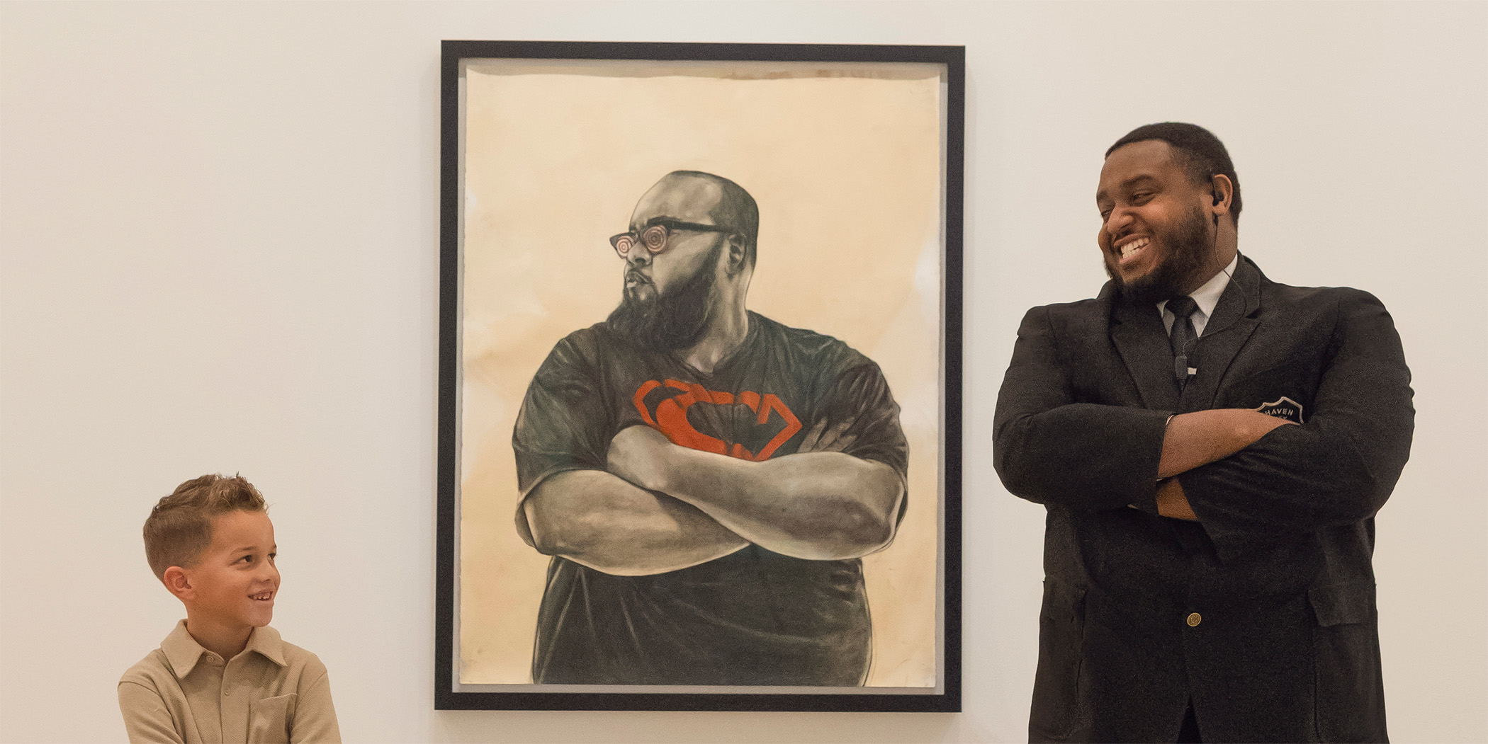 Photo of a young boy and a museum security guard posed smiling with their arms crossed next to a framed drawing of a man with his arms crossed hanging on the wall of an art gallery.