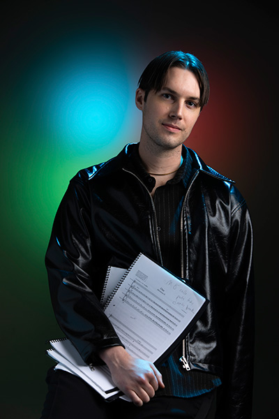 Headshot of a man with brown hair wearing a black leather and black shirt holding notebooks of sheet music.