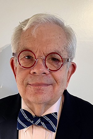 Headshot of a man with light gray hair wearing red framed glasses, a white shirt, a blue and white striped tie, and a navy jacket.