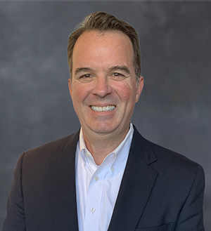 Headshot of a smiling man with short gray hair wearing a white button-down shirt and a dark navy jacket.