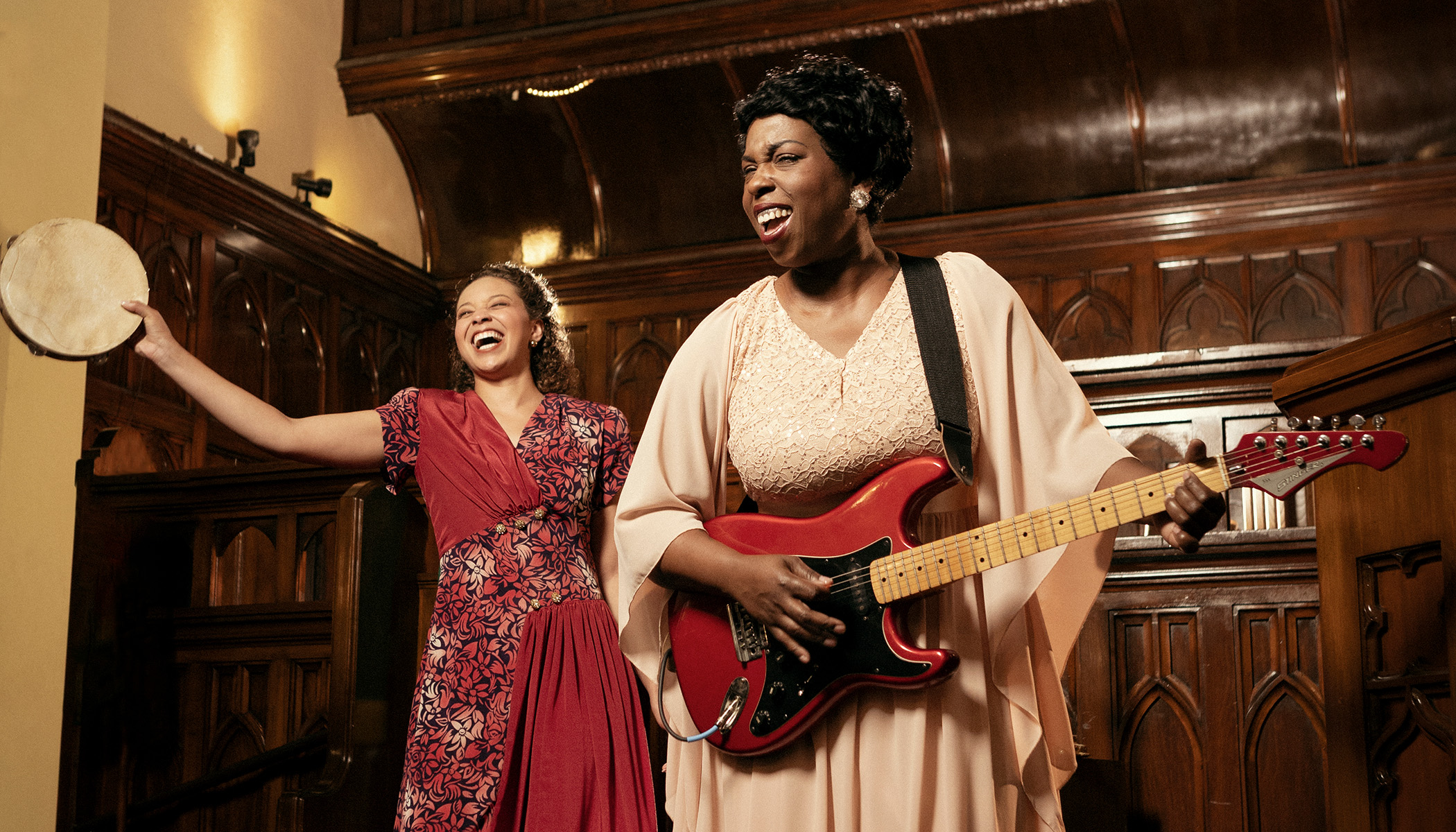 Photo of two women singing in a church. The woman on the left is wearing a red dress and shaking a tambourine. The woman on the right is wearing a pale pink dress and playing a red electric guitar.