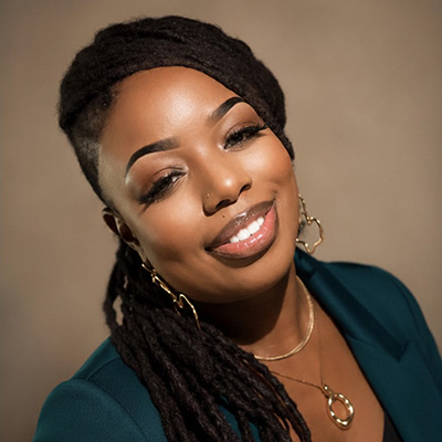 Photo of a smiling woman with long brown braids pulled into a low ponytail. She wears gold hood earrings and necklaces and a dark green blouse.