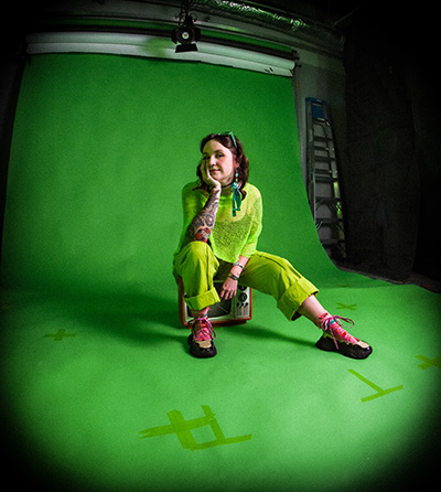 Headshot of Emily Fenton. She has short brown hair and wears a bright green outfit while sitting on a chair in front of a bright green backdrop.