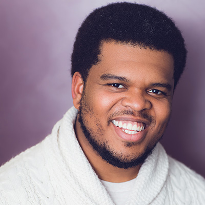 Headshot of Fergie Philippe. He wears a white sweater while smiling in front of a purple background.