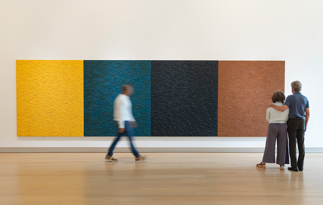 Photo of a man walking in front of a large four-paneled painting on the wall of an art gallery while a man and a woman with their arms around each other stand nearby.
