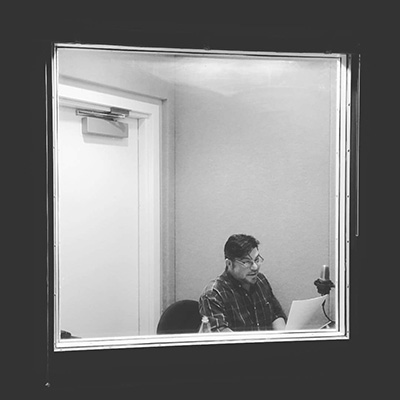 Black and white photo of a man with short dark hair sitting in a sound booth behind a pane of glass and speaking into a microphone. He wears a plaid shirt and glasses.