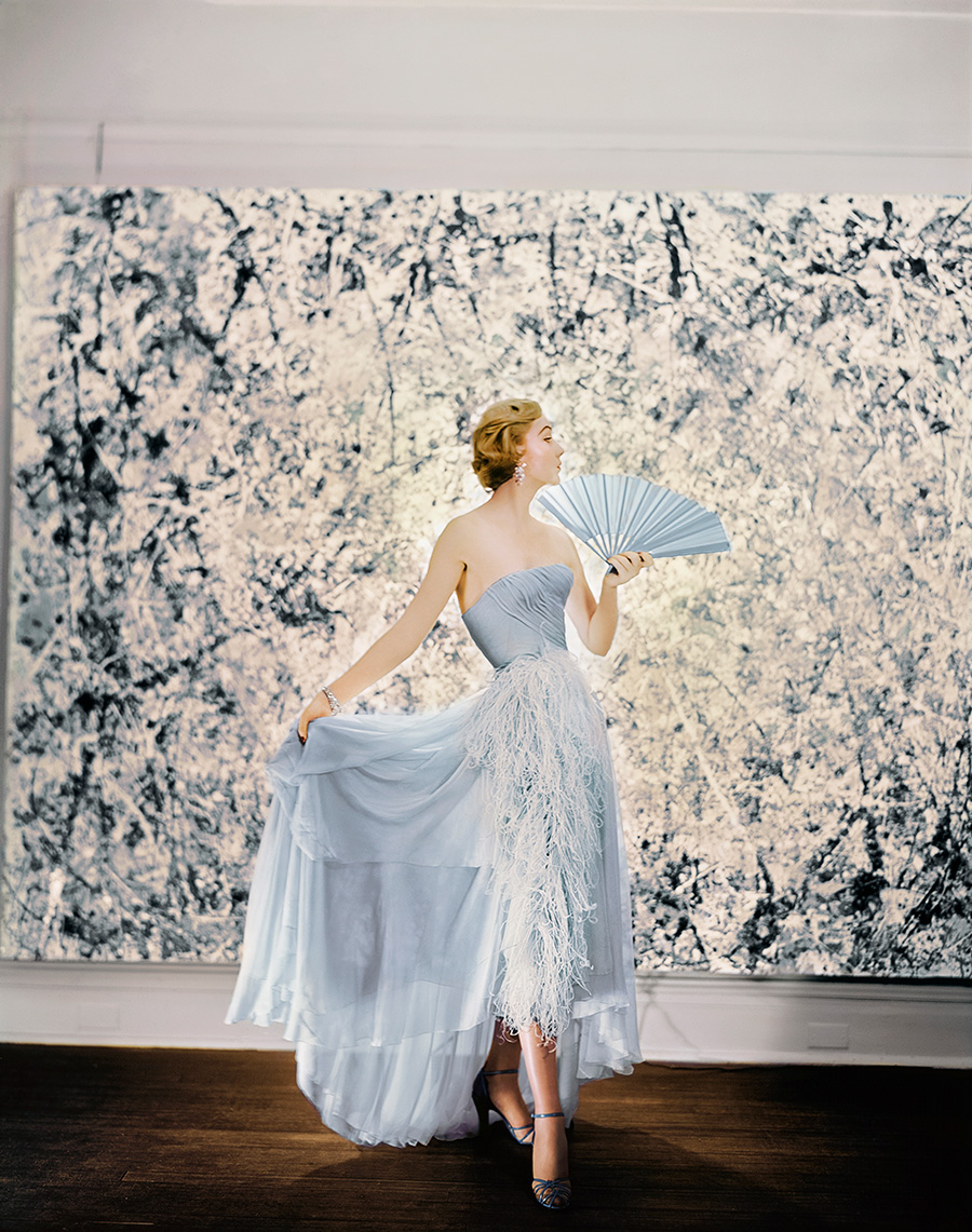 Photo of a woman with short blonde hair posted pearing a light blue evening gown in front of an abstract painting by Jackson Pollock.