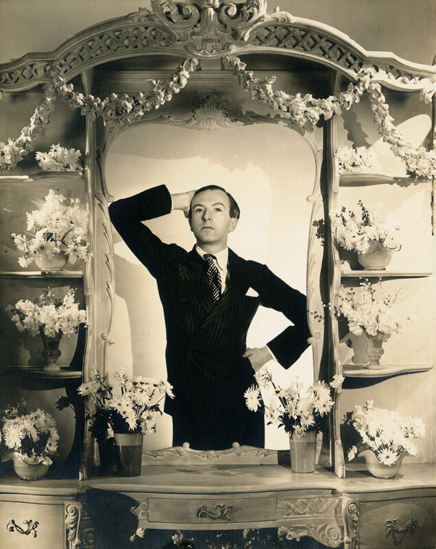 Black and white photo of a man wearing a black suit posing with one arm on his hip and the other on hit head.