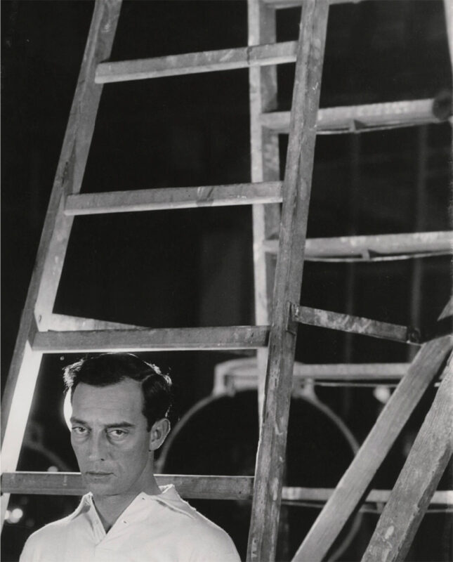 Black and white photo of Buster Keaton standing at the foot of a tall ladder.