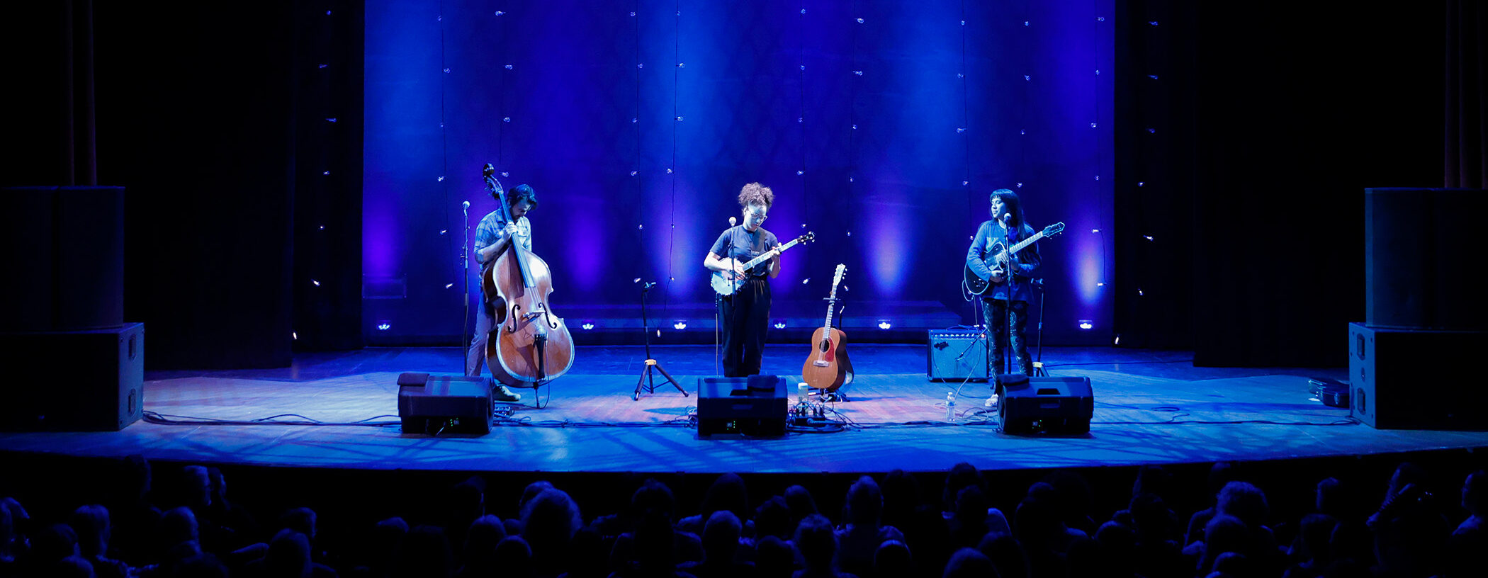 Photo of a man playing an upright bass, a woman playing a banjo, and a woman playing an electric guitar on stage in front of people in seats.