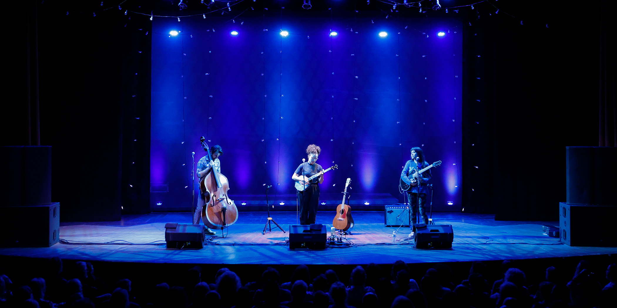 Photo of a man playing an upright bass, a woman playing a banjo, and a woman playing an electric guitar on stage in front of people in seats.
