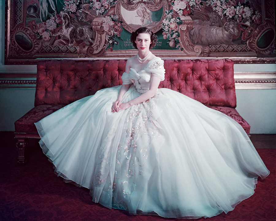 Color photo of a young Princess Margaret wearing a large light blue ballgown and sitting on a tufted red sofa.