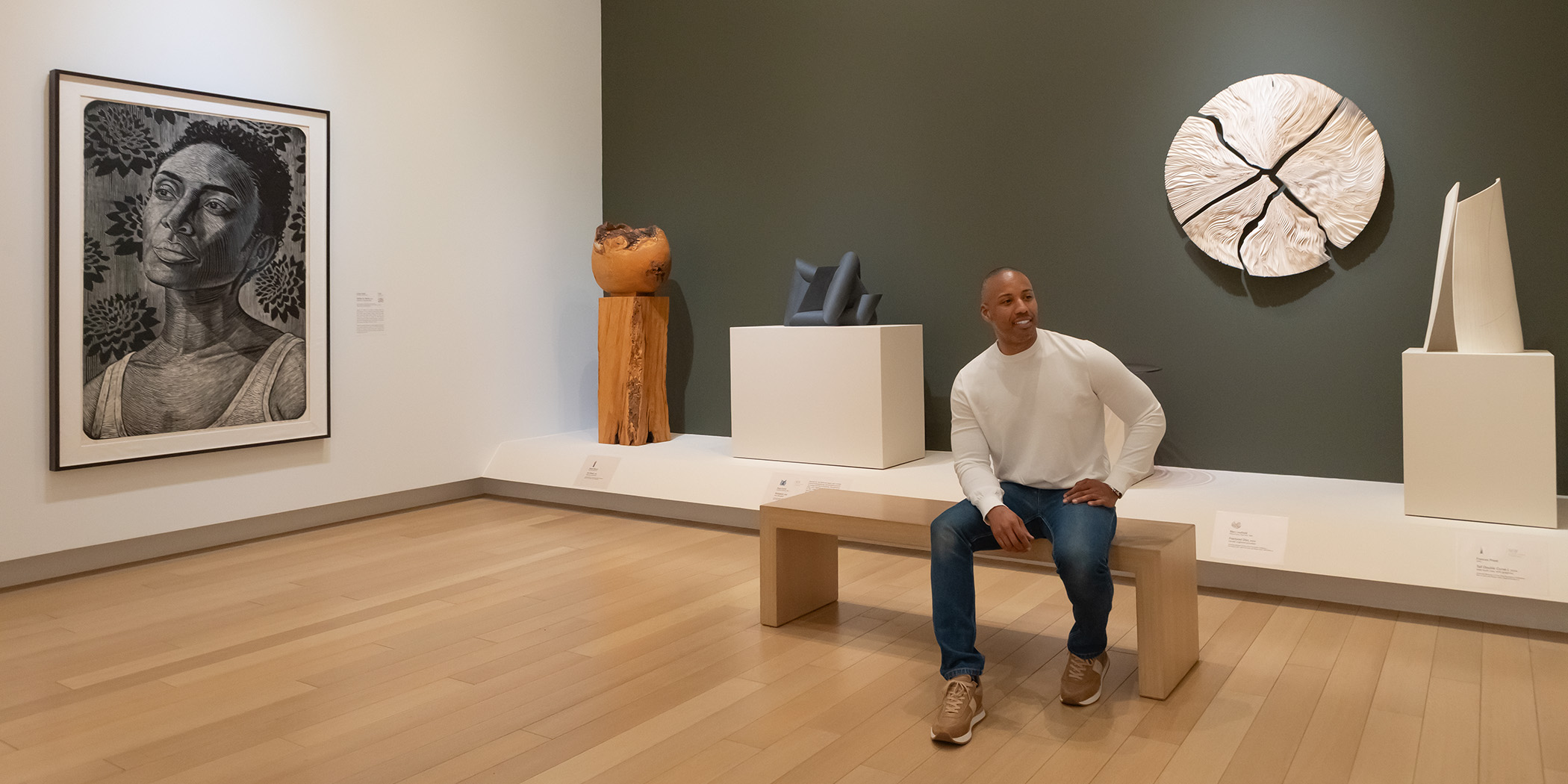 Photo of a man wearing a white sweater and jeans sitting on a bench in an art gallery in front of a wall of sculptures.